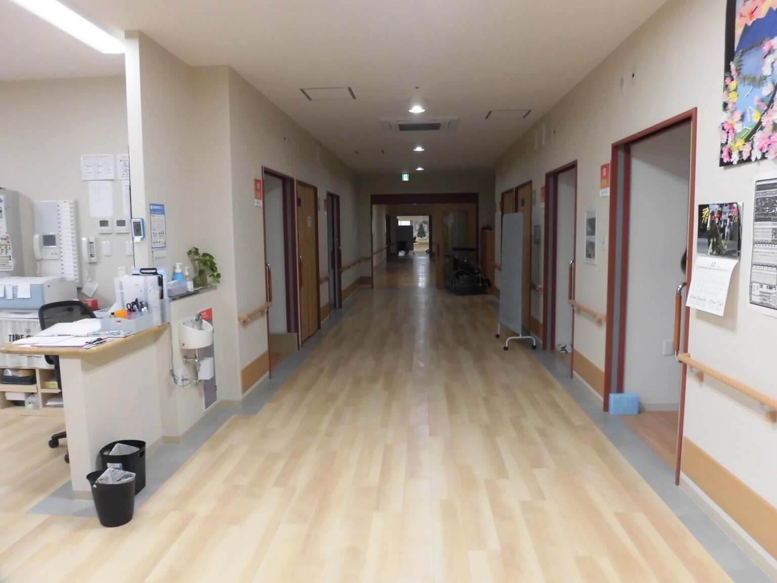 介護施設の廊下。左側に受付カウンター、両側に居室ドア、清潔で明るい空間。